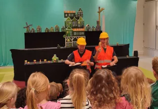 Kinderen kijken naar de voorstelling met op de achtergrond twee acteurs tegen een decor van een stad